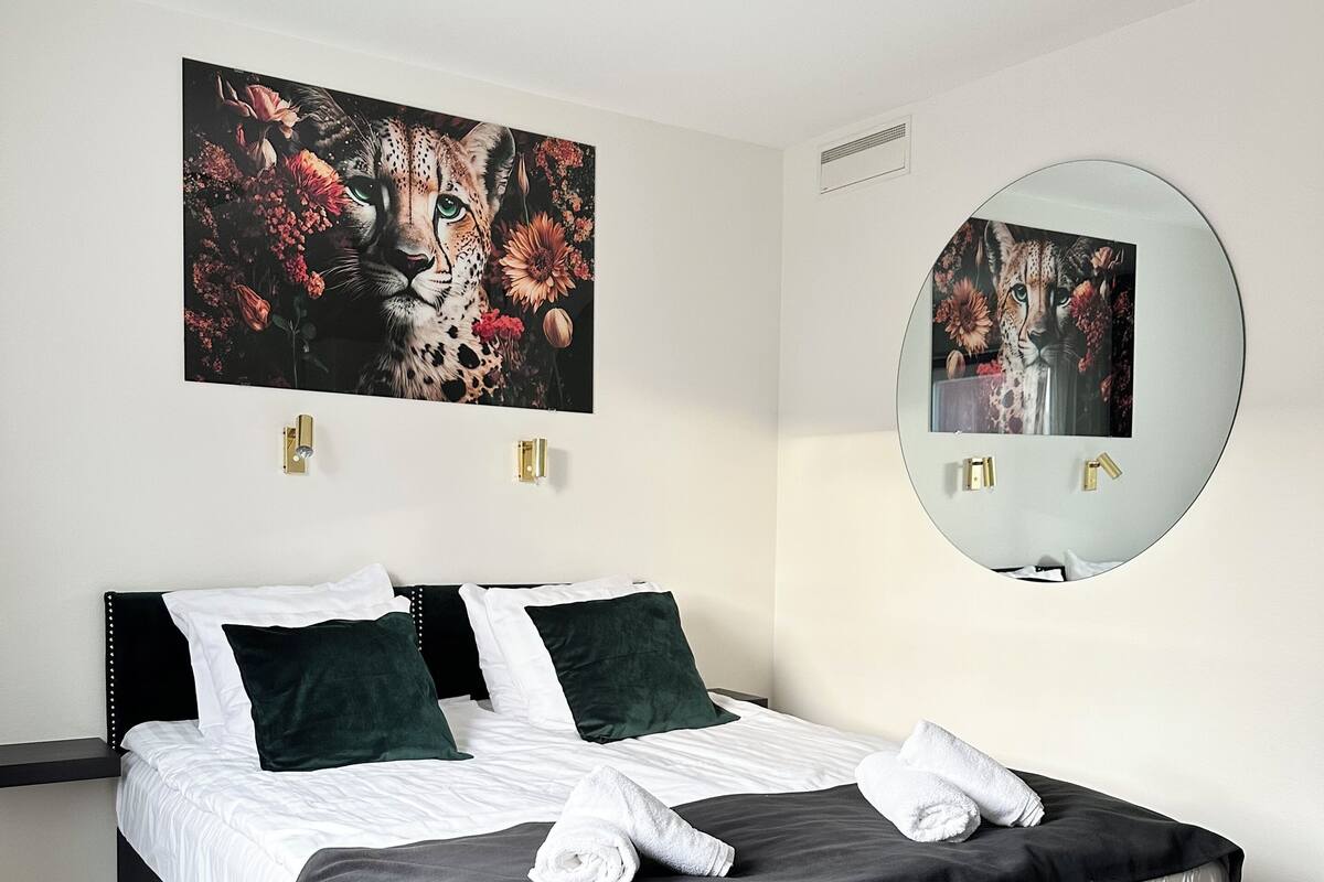 Un lit bien fait, avec une literie blanche et foncée, un grand tableau représentant un léopard, un miroir rond et un luminaire fixé au mur.