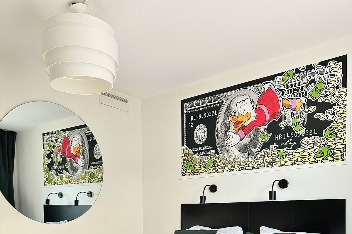 Un lit bien fait, avec du linge de lit blanc et des coussins verts, un grand miroir rond accroché au mur et une grande affiche représentant un personnage de dessin animé entouré d’argent.