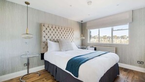 Une chambre moderne avec une tête de lit capitonnée, une table de chevet ronde, une grande fenêtre donnant sur la ville et un plaid bleu sur le lit.