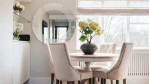 Un coin repas avec une table ronde, quatre chaises rembourrées, un vase de fleurs, un miroir rond et une fenêtre donnant sur des arbres.