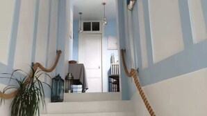 Un escalier avec des marches blanches et des murs rayés de bleu et de blanc. Il y a un pot de plante sur la gauche et une main courante en corde sur la droite.