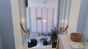 Un couloir étroit avec un escalier menant à une pièce où se trouvent un lit, une table avec un panier et une plante en pot.
