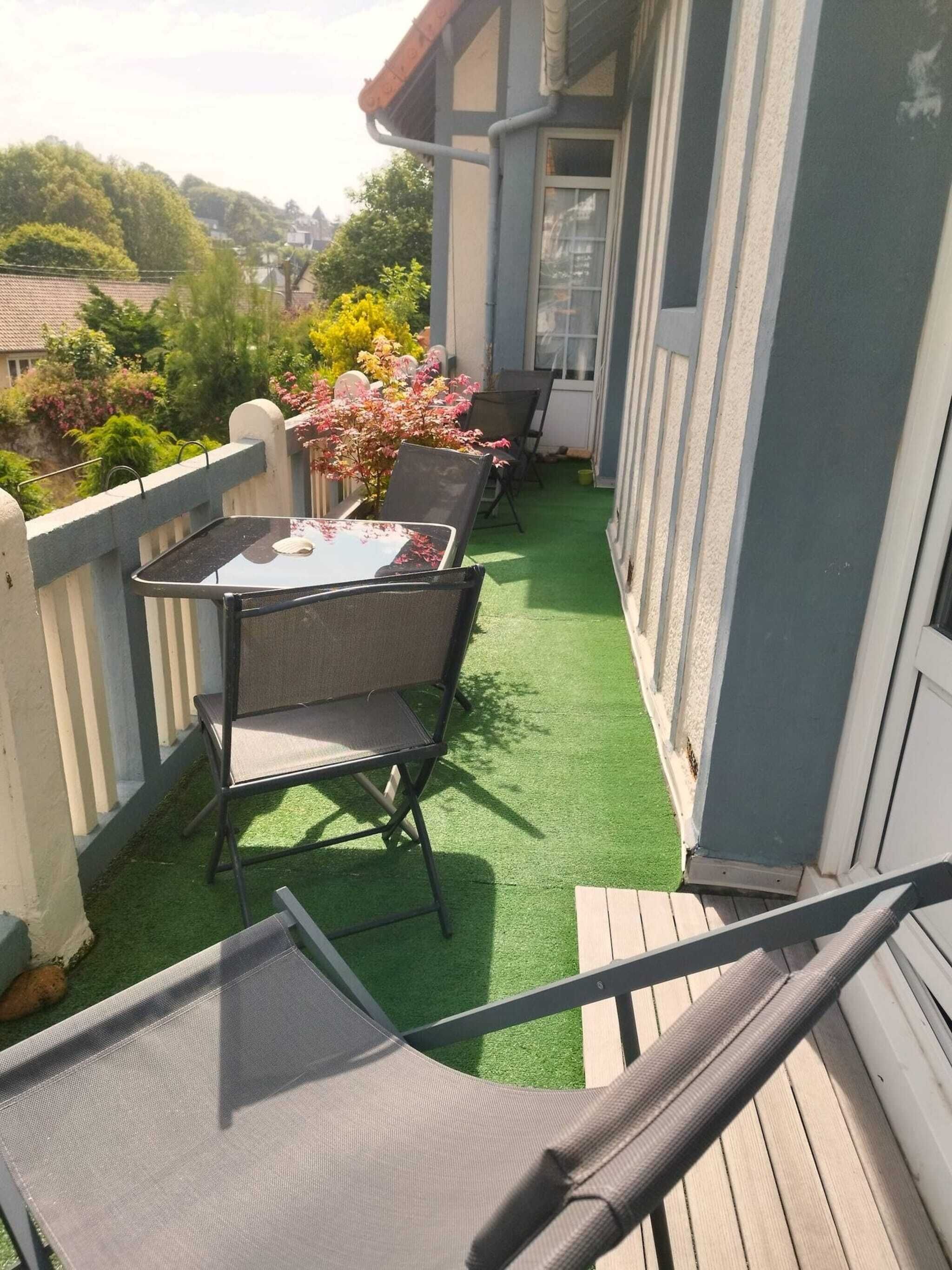 Ein Balkon mit kleinem Tisch und zwei Stühlen, umgeben von einer Holzterrasse und einer Grünfläche mit künstlichem Rasen.