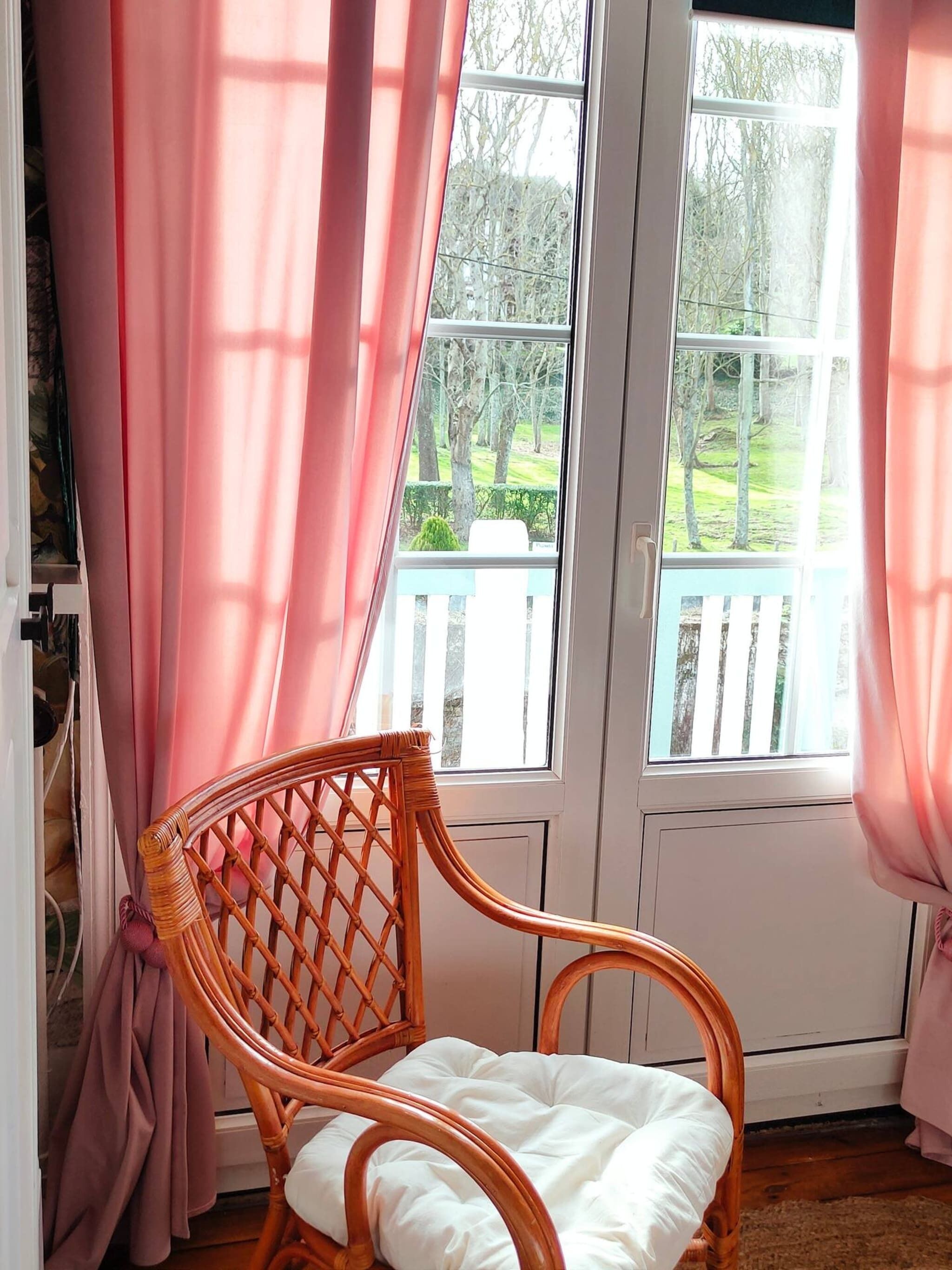 A wicker chair with a white cushion in a room with pink curtains and a view of a garden through a white door.