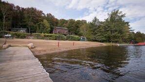 A wooden dock extending into a calm lake, surrounded by trees and a building with a sloped roof.