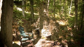 A rustic stone fireplace surrounded by trees, with a green Adirondack chair and a fire pit nearby.