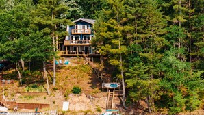 A lakeside cabin with a deck, surrounded by dense trees and docked boats. - Lake View & Access/Hot Tub/5000 Arcade Games/Pool Table/Pontoon for Rent (Kalkaska)