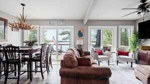 A spacious living room with a brown leather sofa, a dining area with wooden chairs, and a chandelier. - Lake View & Access/Hot Tub/5000 Arcade Games/Pool Table/Pontoon for Rent (Kalkaska)
