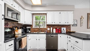 A modern kitchen with white cabinets, a black countertop, a stainless steel dishwasher, and a brick backsplash. - Lake View & Access/Hot Tub/5000 Arcade Games/Pool Table/Pontoon for Rent (Kalkaska)