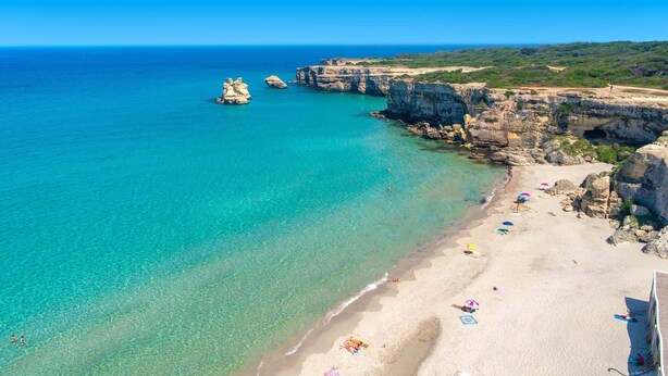 Ein Küstenstrand mit klarem türkisblauem Wasser, felsigen Klippen und einem sandigen Ufer mit Sonnenschirmen und Badegästen.