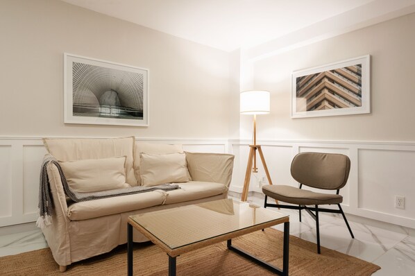 Un salon moderne avec un canapé beige, une table basse en verre, un lampadaire et deux œuvres d’art encadrées accrochées aux murs.