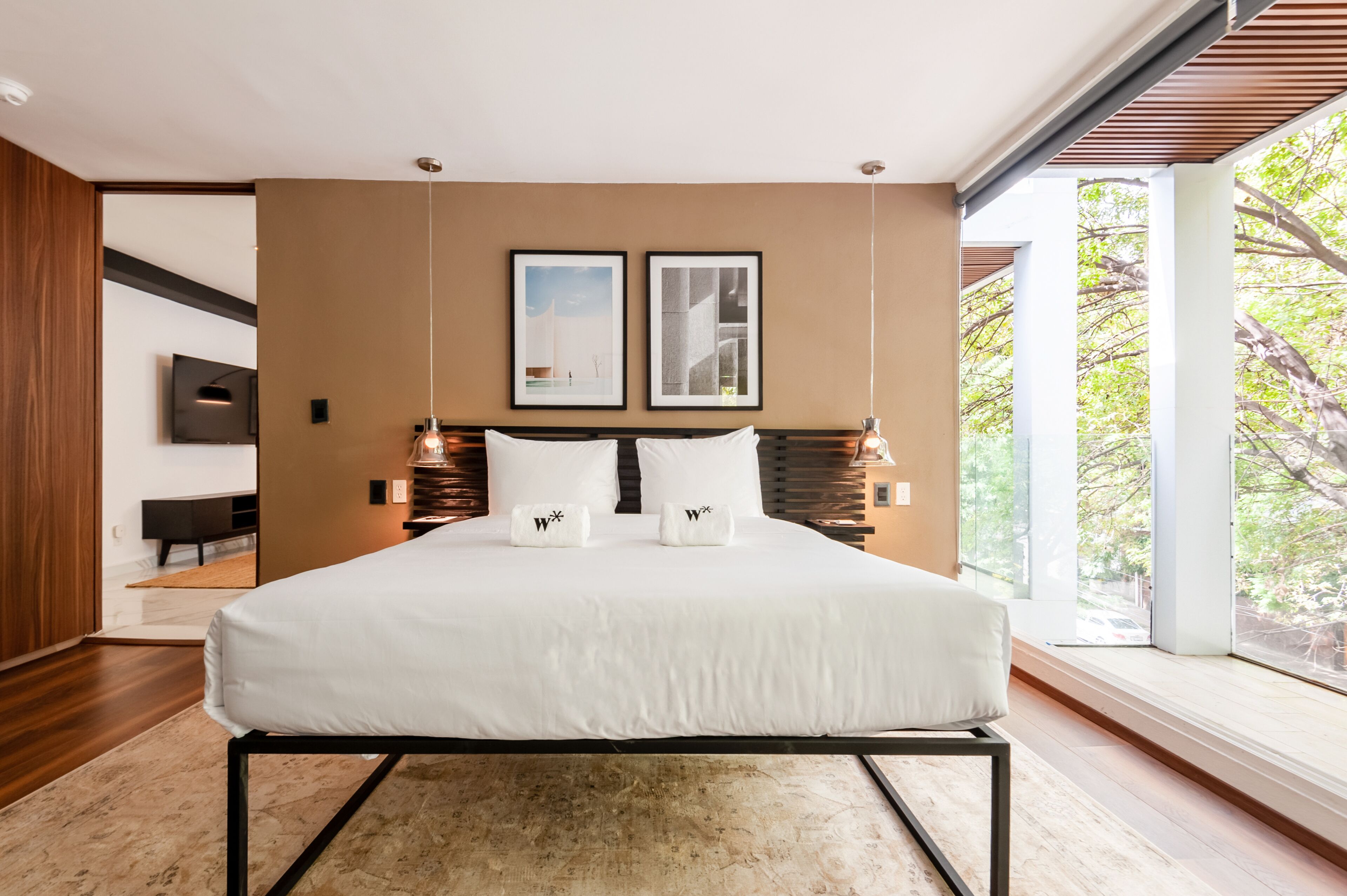 Une chambre moderne avec un grand lit, deux tableaux encadrés au mur et une vue sur les arbres à travers de grandes fenêtres.