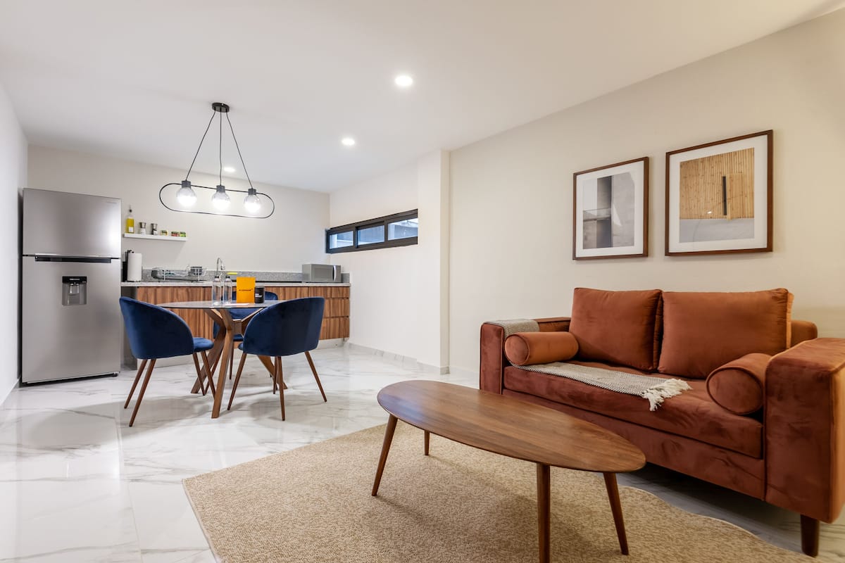 Una cocina moderna con refrigerador, microondas y gabinetes de madera. Hay un comedor con una mesa y sillas, y una sala de estar con sofá y una mesita de centro.