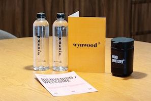 Two bottles of Wynwood House water, a black hand sanitizer, a yellow box, and a welcome card on a wooden table.