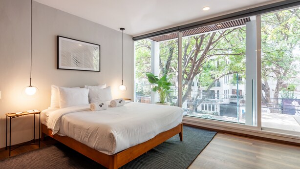 A modern bedroom with a large bed, bedside tables, a framed artwork, and a view of trees and buildings through large windows. - Wynwood House Allan Poe (Mexico City)