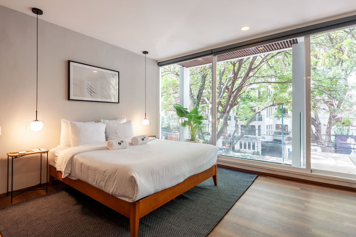 Un dormitorio moderno con una cama grande, mesitas de noche, una obra de arte enmarcada y vistas a árboles y edificios a través de amplios ventanales.
