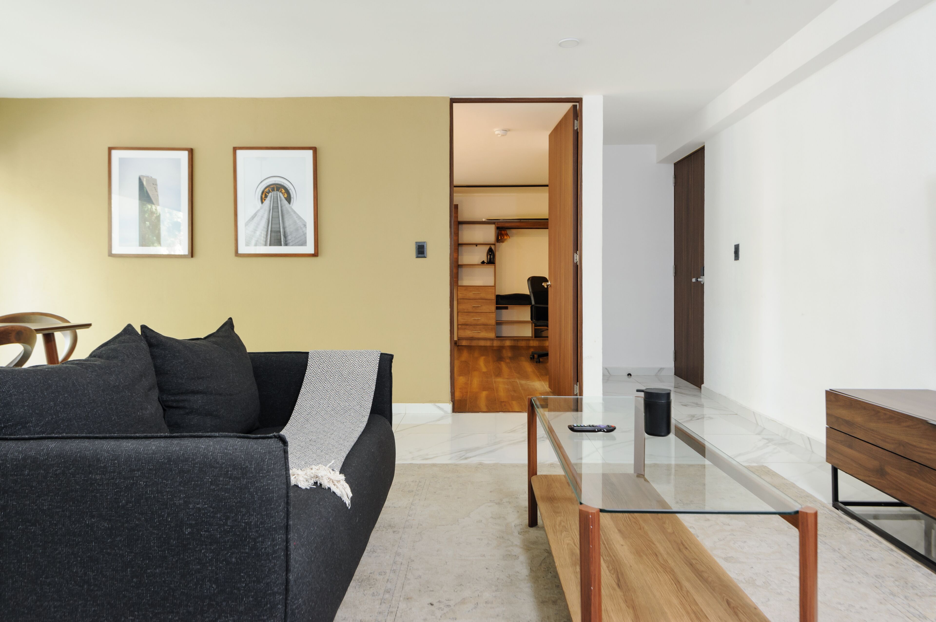 Un salon moderne avec un canapé, une table basse en verre et une table d’appoint en bois. Deux tableaux encadrés sont accrochés au mur.