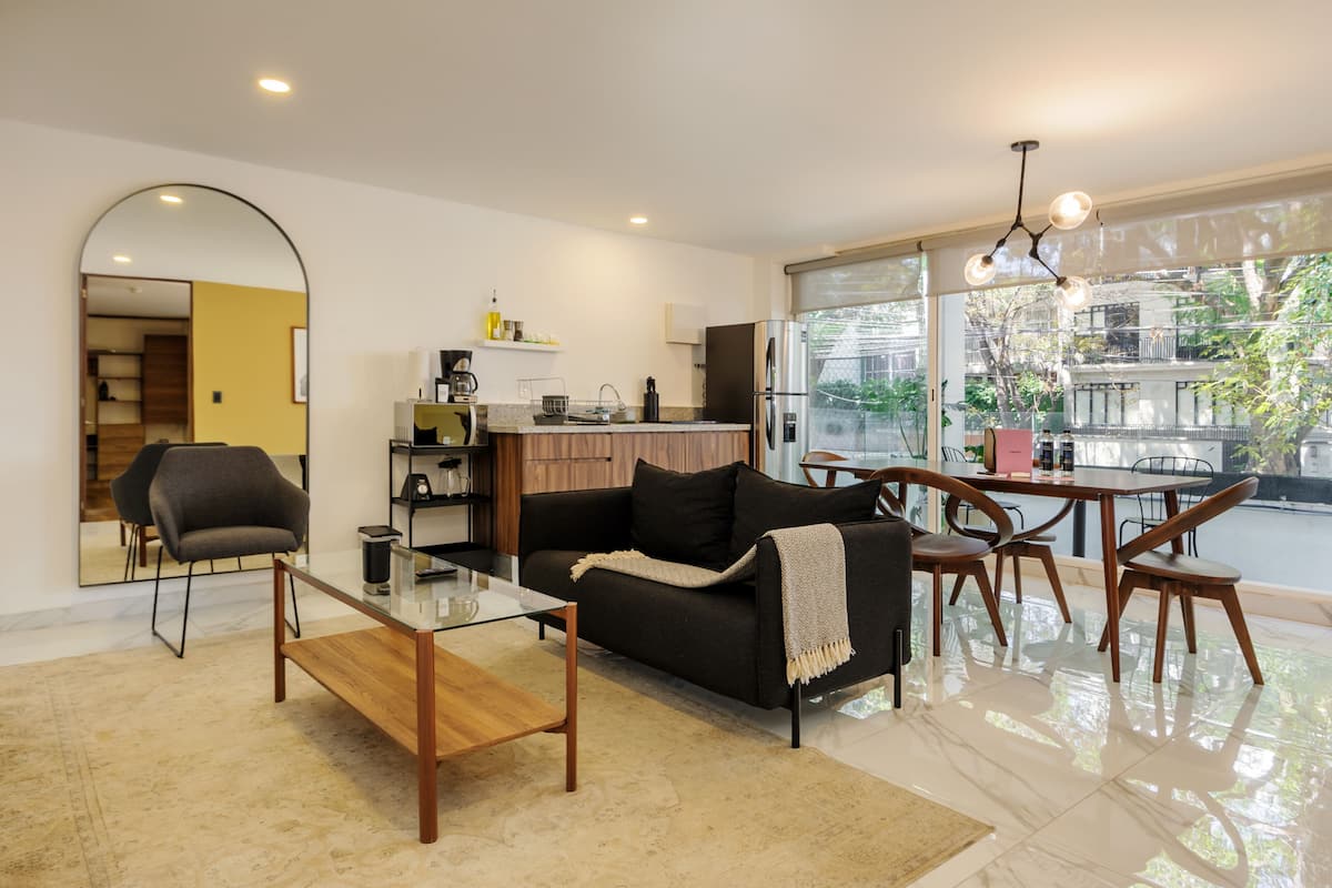 Una sala de estar moderna con una mesa de centro de vidrio, un sofá negro y un comedor con sillas de madera.