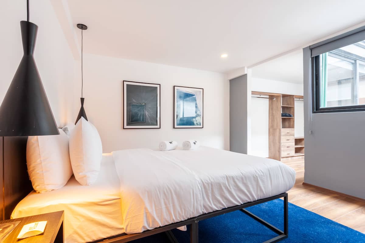 Un dormitorio moderno con cama, ropa de cama blanca, una lámpara colgante negra y dos cuadros en la pared.