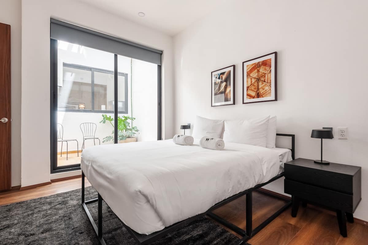 Un dormitorio moderno con una cama grande, una mesita de noche y un ventanal corredero que da a un balcón con sillas.