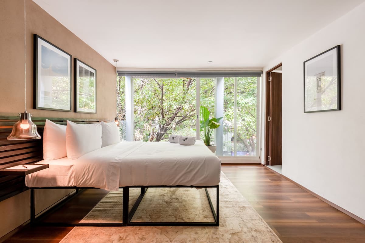 Un dormitorio moderno con una cama grande, una mesita de noche, una lámpara y vistas a zonas verdes a través de amplios ventanales.