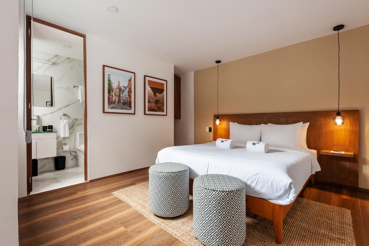 Una habitación de hotel moderna con una cama grande, dos otomanas, un cabecero de madera y cuadros enmarcados en la pared.
