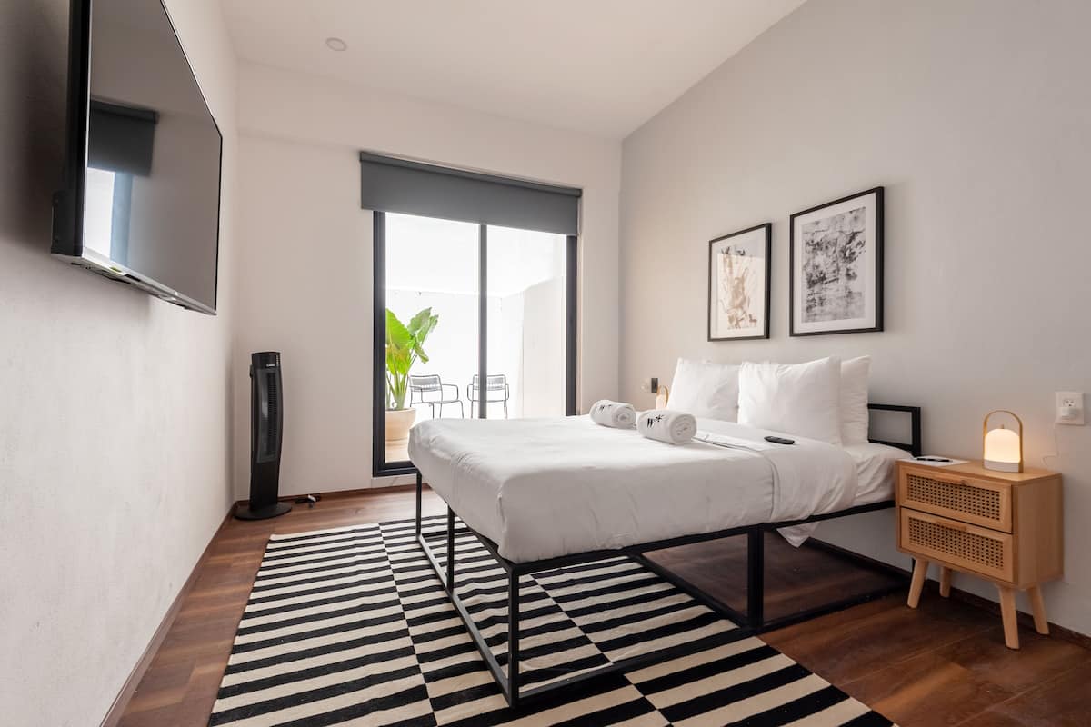 Un dormitorio moderno con una cama, un ventilador, una mesita de noche con una lámpara y dos cuadros enmarcados en la pared.