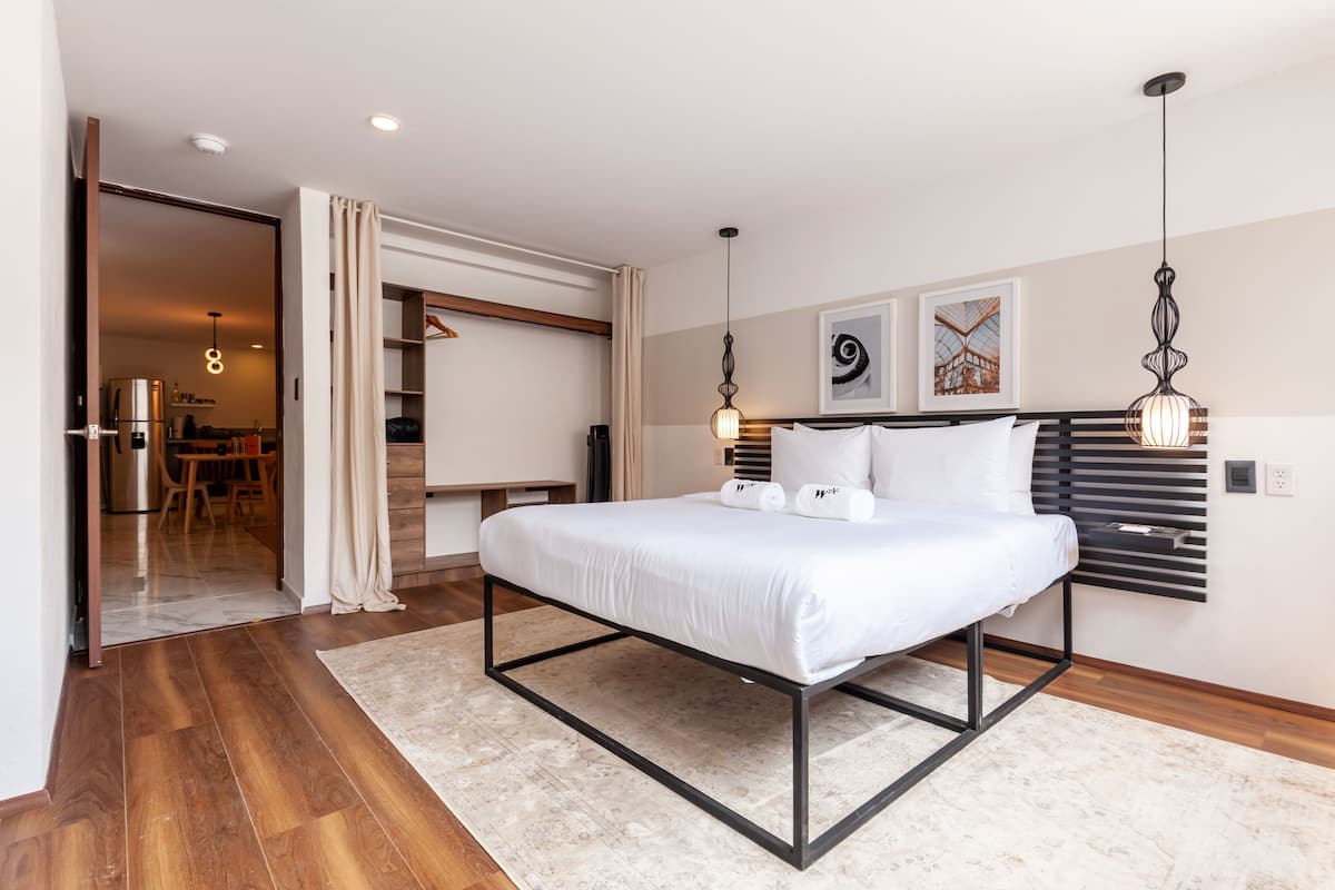 Un dormitorio moderno con cama, mesitas de noche, armario y un área de comedor visible a través de una puerta abierta.