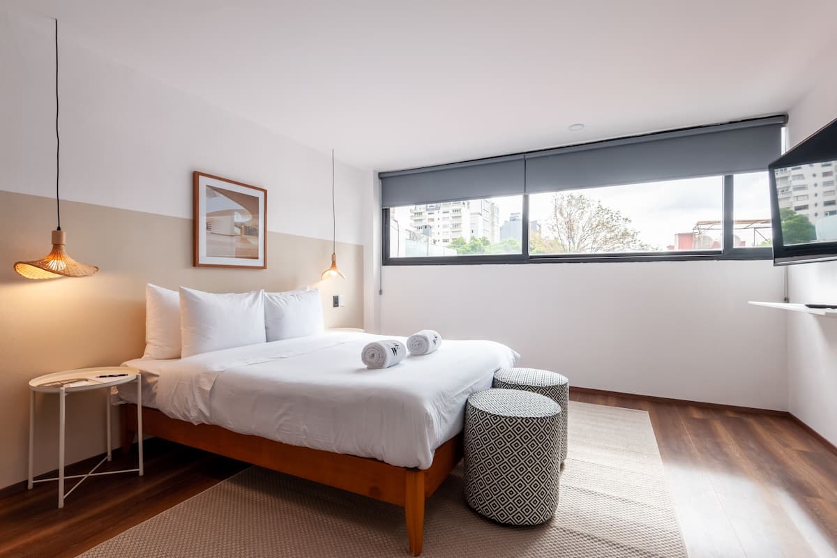 Un dormitorio moderno con una cama de madera, ropa de cama blanca, un cuadro en la pared, un ventanal grande y una pequeña mesa redonda.