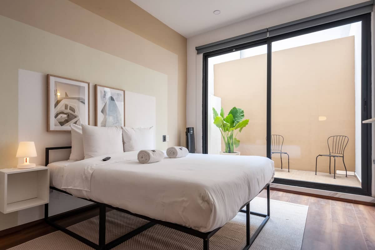 Habitación de hotel moderna con una cama grande, una mesita de noche con lámpara, una planta en maceta y vistas al balcón exterior.