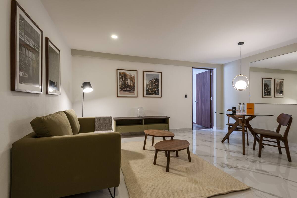 Una sala de estar moderna con sofá, mesa de centro y comedor con sillas. Hay cuadros enmarcados en las paredes y una lámpara en el suelo.