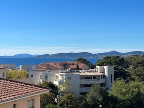 Una zona residencial costera con edificios, árboles y una vista despejada del mar y colinas distantes.