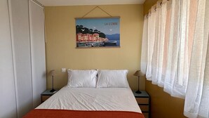 Une chambre à coucher comprenant un lit, deux tables de chevet, une commode, un tableau accroché au mur et une fenêtre avec des rideaux.