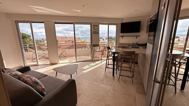 Un vaste espace de vie comprenant un canapé, une petite table et un coin bar avec des tabourets. De grandes portes-fenêtres coulissantes donnent accès à un patio extérieur avec des chaises et une table.