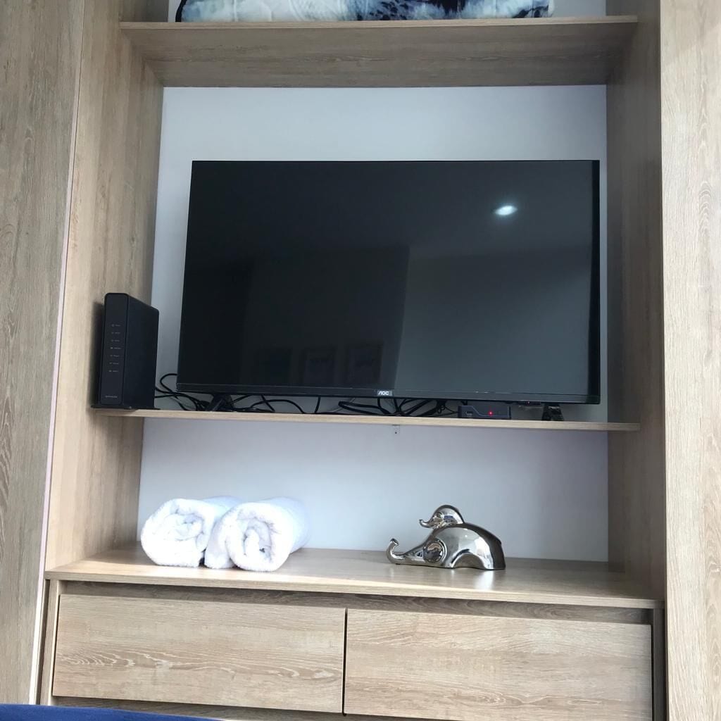 Un mueble bajo de madera para televisor con un televisor de pantalla plana, un dispositivo electrónico negro y dos toallas enrolladas en el estante inferior.