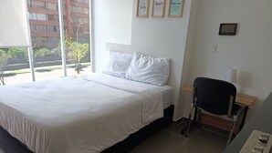 A modern hotel room with a large bed, a desk with a chair, and a window offering a view of the outside. - Loft Comodo, Iluminado en Usaquen (Bogotá)