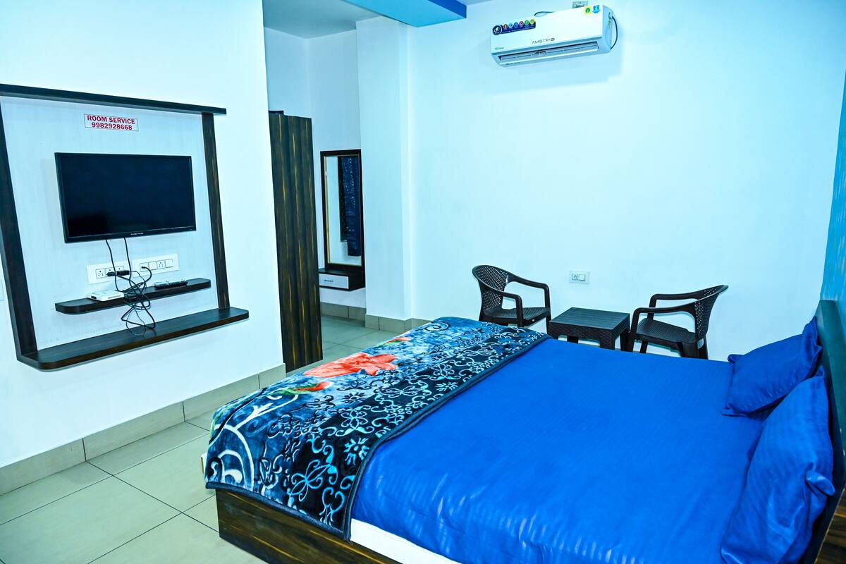 A hotel room with a bed, a TV mounted on the wall, a desk with a chair, and an air conditioning unit.