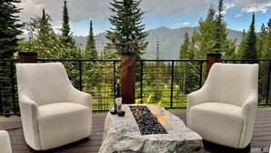 A deck with two white armchairs facing a stone fire feature and a bottle of wine, surrounded by a metal railing with a scenic mountain view. - Mountain Top Serenity (Big Sky)