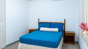 Cama bem arrumada com colcha azul e travesseiros brancos em quarto com paredes brancas e cabeceira de madeira.