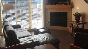 Ein Wohnzimmer mit einer Couch, einem an der Wand befestigten Fernseher, einem Kamin und einem großen Fenster mit Jalousien.