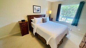 A bedroom with a large bed, a nightstand, a bedside lamp, a framed picture on the wall, and a window with curtains. - Ocean View Townhome - Sooke Harbour Marina (Sooke)