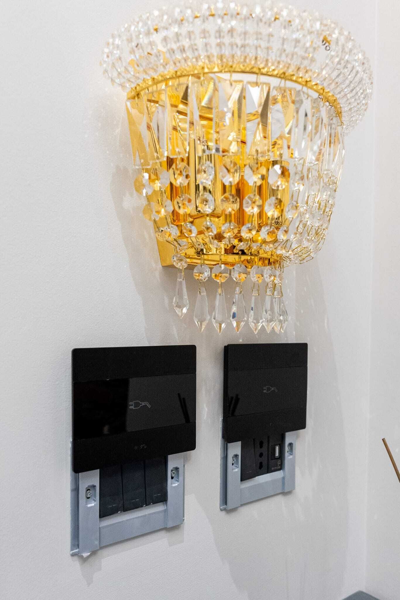 A chandelier with a gold frame and crystal decorations, mounted on a white wall. Below it, there are two black electrical switches.