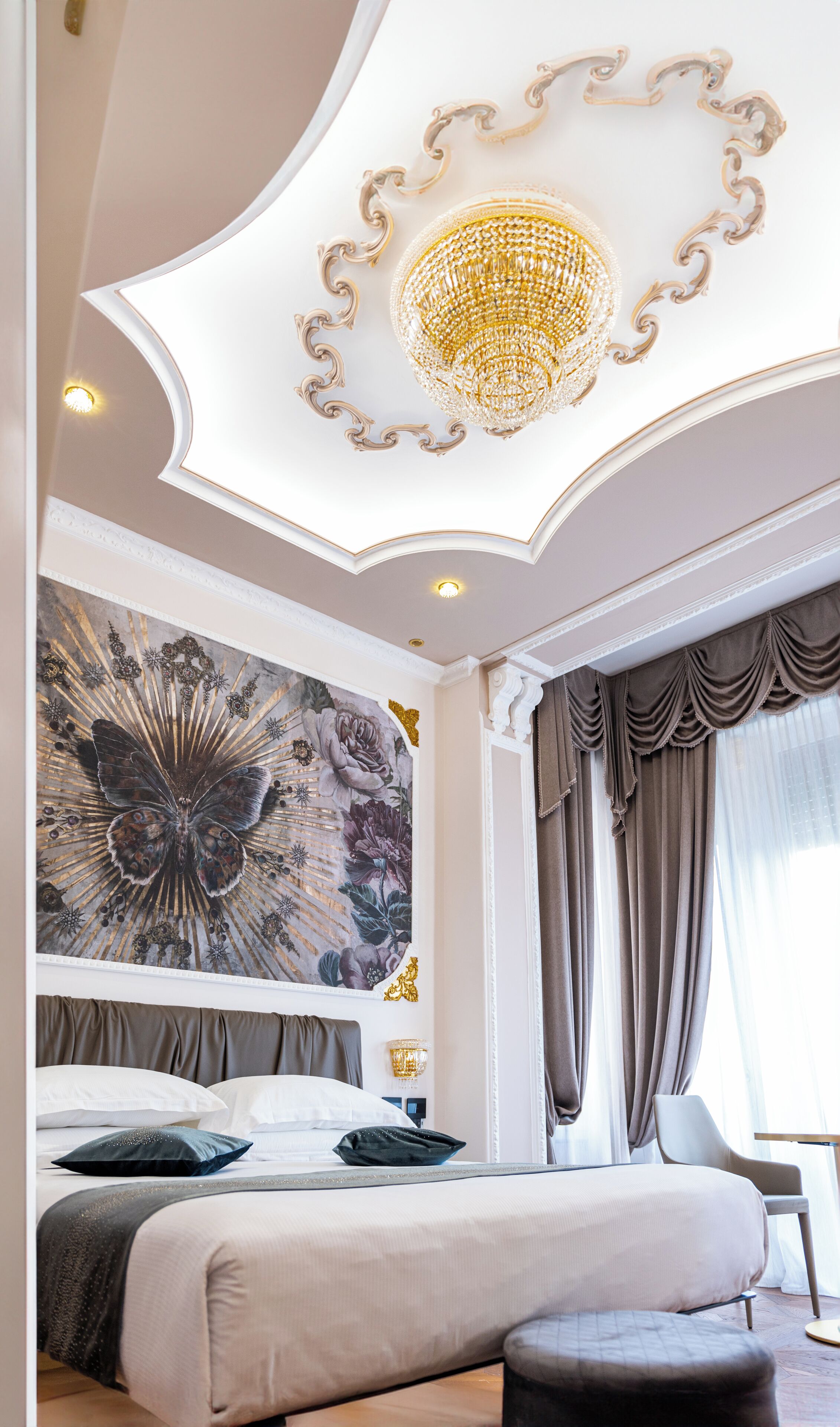 Une chambre à coucher luxueuse, dotée d’un grand lit, d’un lustre, d’une fresque florale et offrant une vue sur l’extérieur.