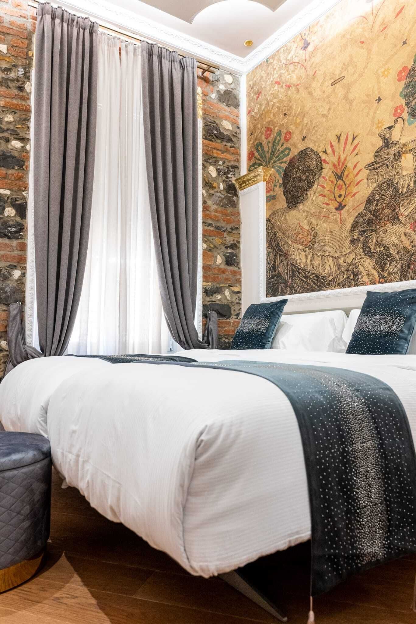 A bedroom with a large bed, grey curtains, a quilted ottoman, and a mural on the wall.