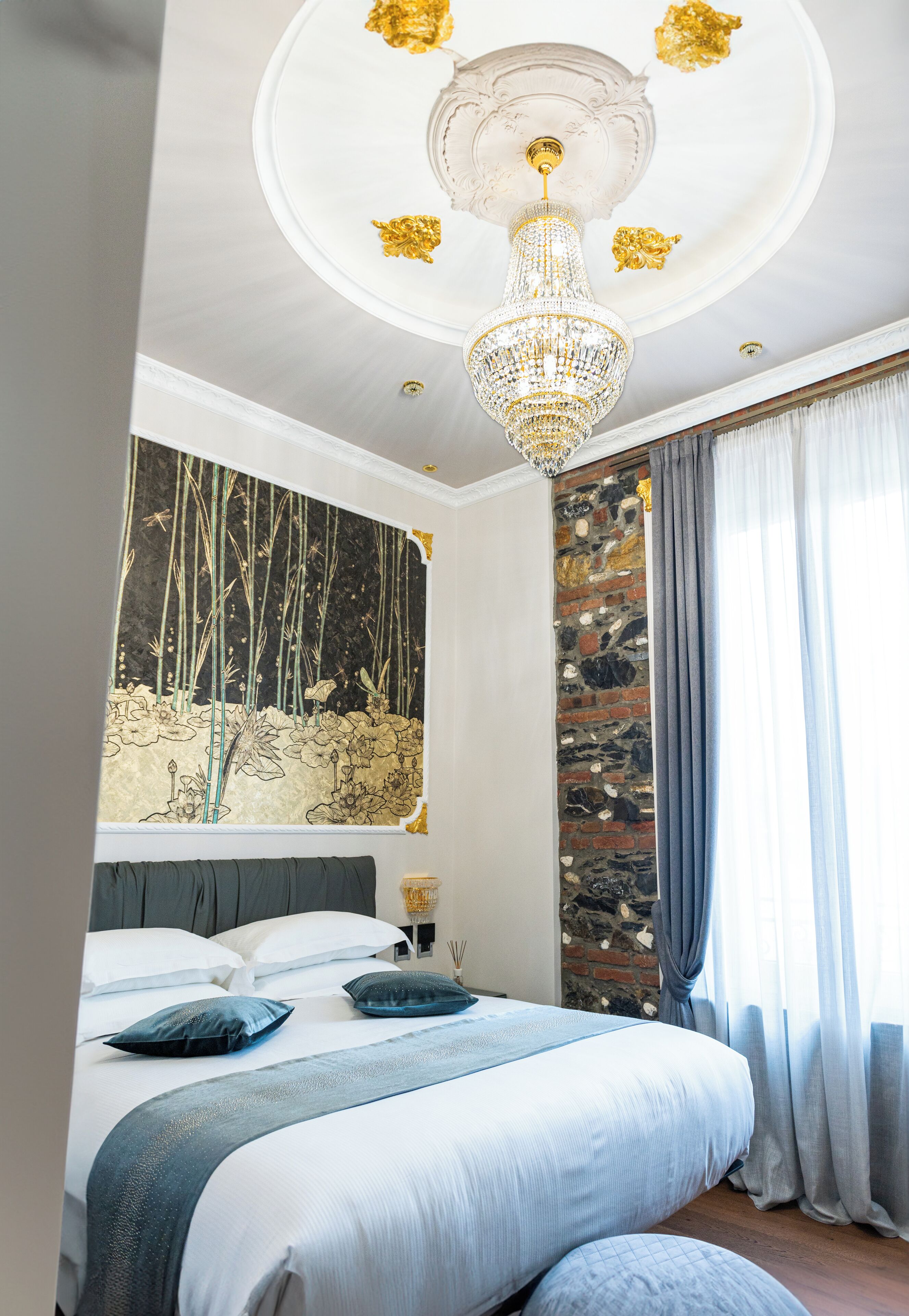 Une chambre avec un grand lit, un lustre, un mur orné d’une fresque représentant une forêt de bambous et un mur en pierre.