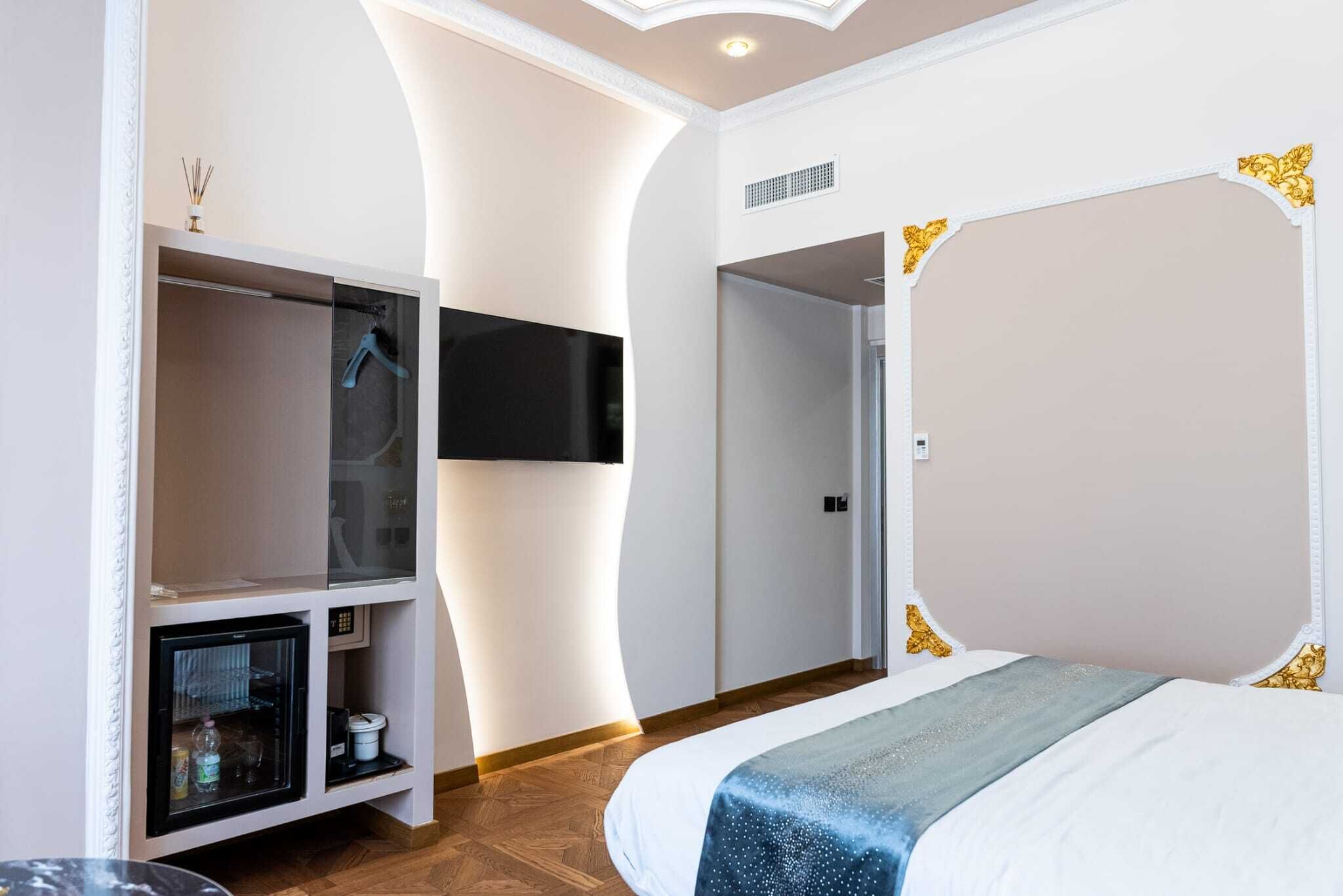 A modern hotel room with a curved ceiling, a flat-screen TV mounted on the wall, a bed with a blue and white bedspread, and a built-in wardrobe with a mirror and shelves.