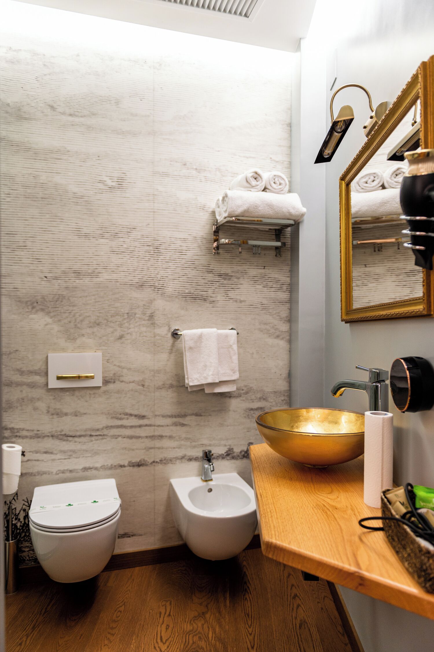 A bathroom with a wooden vanity, a gold sink, a bidet, and a towel rack with towels.