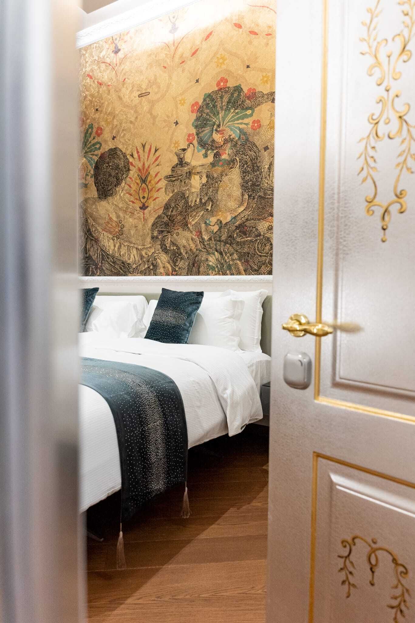 A bedroom with a bed, white bedding, a dark blue throw, and a wall with a floral tapestry.