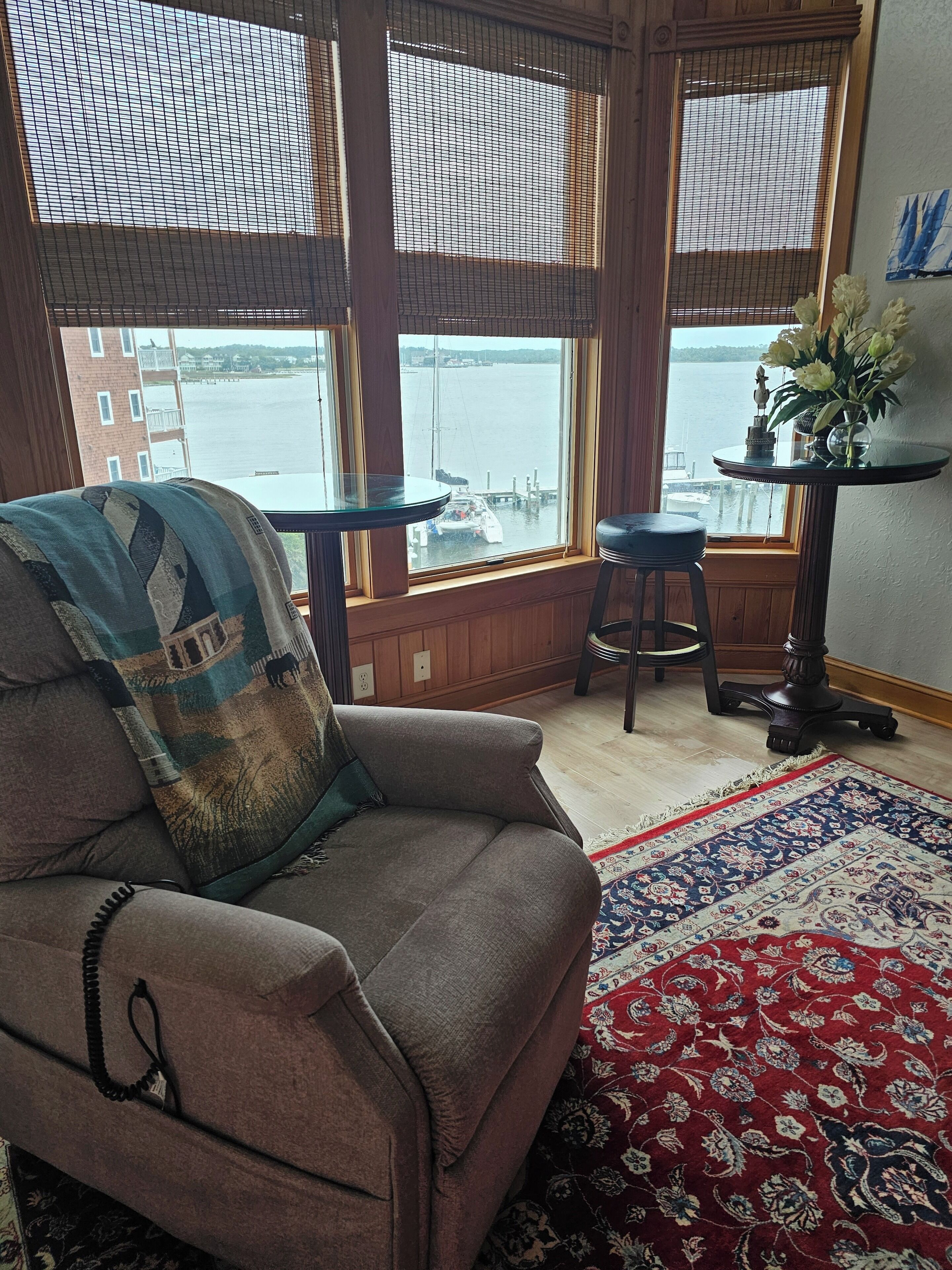 Un cuarto acogedor con un ventanal grande que da a una extensión de agua, un sillón cómodo, una mesita redonda con un florero y una alfombra estampada.