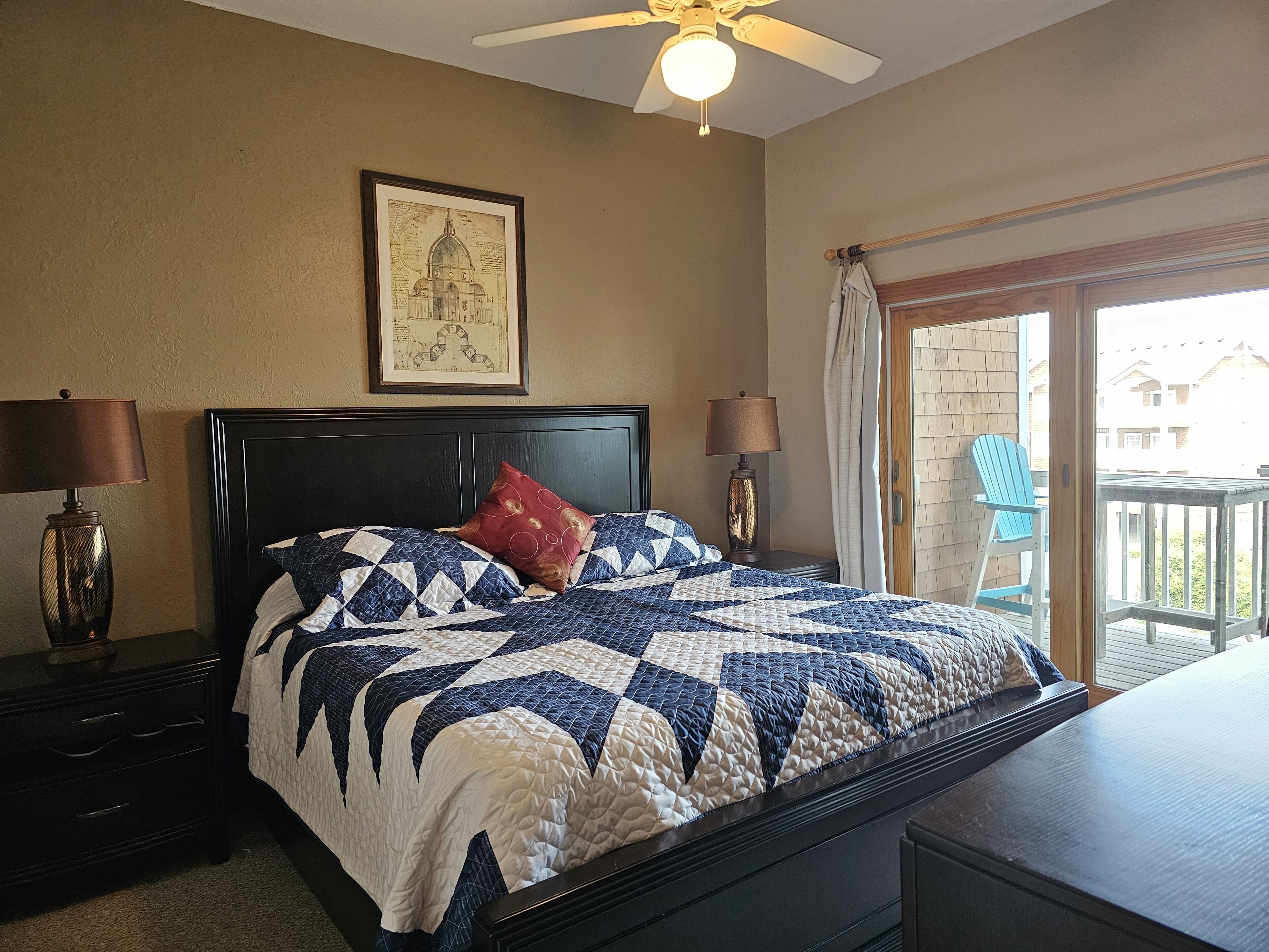 A bedroom with a large bed, two bedside tables with lamps, a ceiling fan, a framed map on the wall, and a view of the outside balcony.